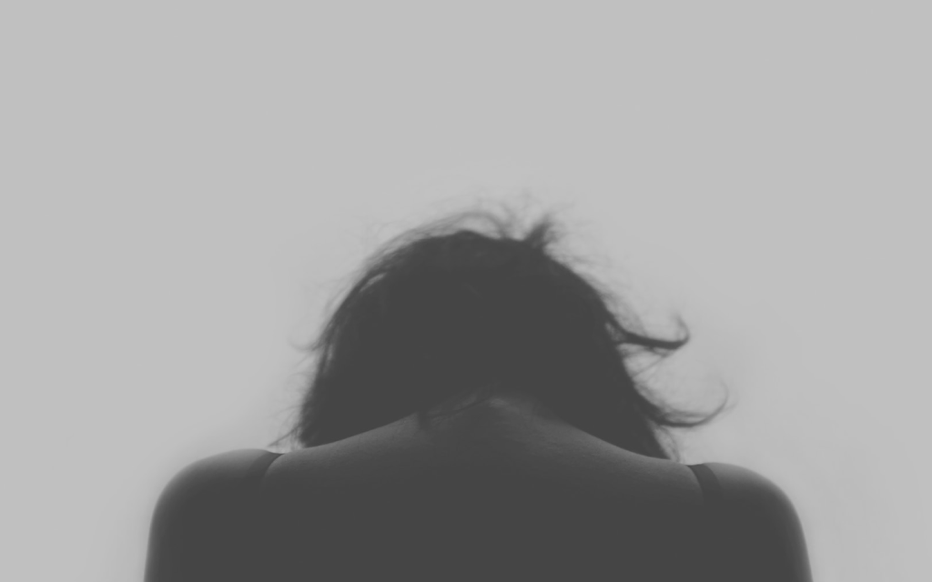 This is a black and white image of a White woman with dark hair with her head down away from the camera. The woman is wearly a black camousel so that the top third of her back and shoulders is perceptible. With her head slightly bowed, she seems sad, much like the general energy of Oh Jin-Hee in "Emergency Couple" because of her toxic marriage.