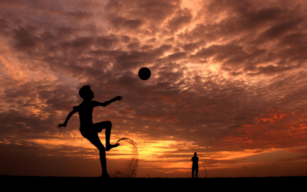 This is a shoeless kid mid kick of a foot ball and a figure of an adult in the background watching. The sky is orange, and greyish, almost like flames. It represent the power of soccer in "The English Game".