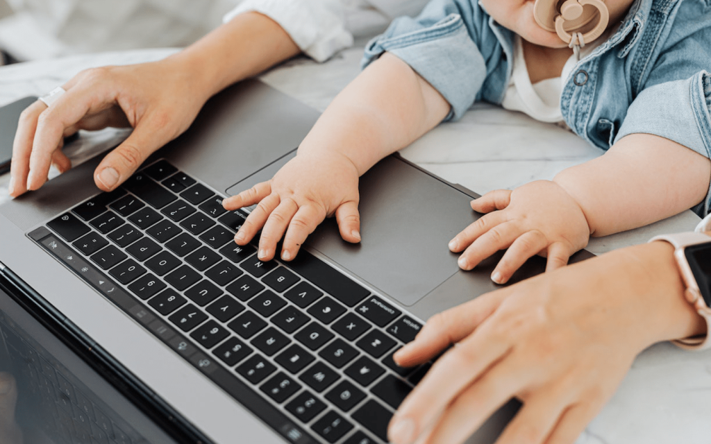 This is a-person-of a woman's hands using-a-laptop with the hands of an infant also on the laptop. This represent the workin mom.