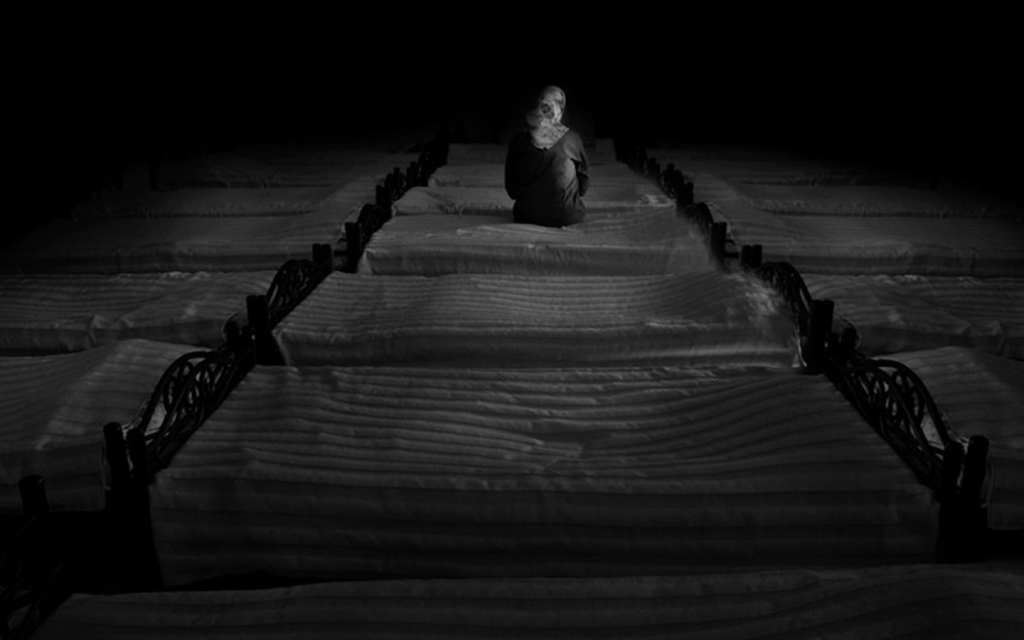 This is a black and white image of the back of a woman with a head scarf, sitting on a bed in a dark room with a row of beds; she is alone in the room. This is used to represent the sullen atmosphere of Killing Eve in season 3 episode 5 as we learn about Villanelle's history.