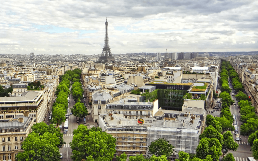 This is two branched streets of the City of Paris lined with trees. The Eifle tower is shown against a slightly cloudy but bright greyish sky. The are building between the two streets and cars in the streets. This is used to represent Emily's view of Paris in "Emily in Paris".