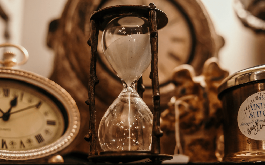 This is a dark wooden hourglasson a table with other items against the back drop with a larger clock. There is also an aged golden clock at the corner left, and a borwn candle case with a golden lid. It is used to signify the time aspect of the "About Time" movie.