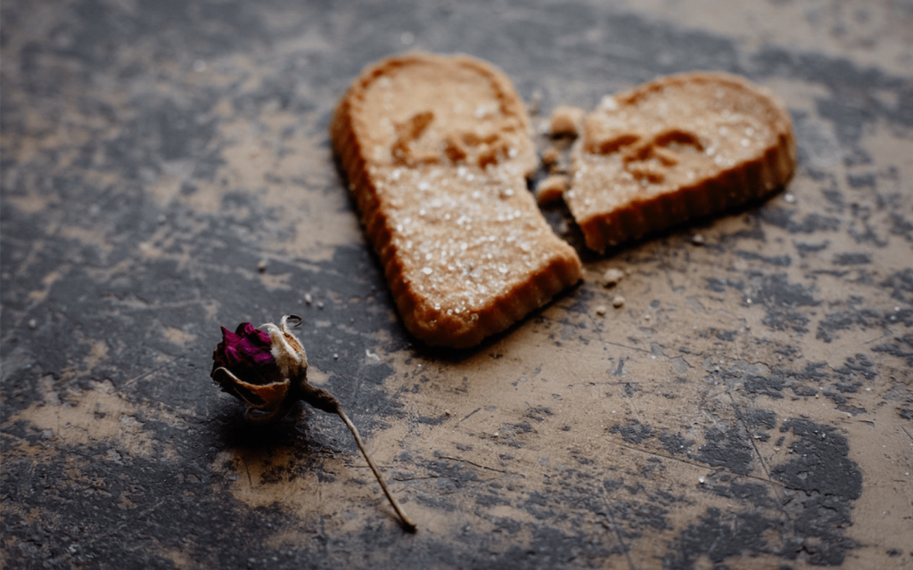 This is a broken heart shaped ginger bread cookie with the words 'LOVE' inscribed on top. The cookie is against a rough back drop and has been broken but piece back together. There is also a dying red rose near the bottom of the cookie. This is used to represent heartbreak in Season 3 episode 3 of "Grown-ish".